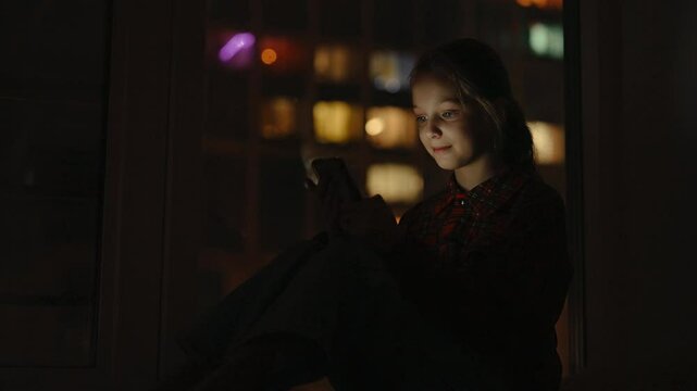 Happy 10 yo child girl smiling looking to screen while typing on smartphone, sitting alone at night by window colorful bokeh lighting creating vibrant nighttime ambiance on blurred background.