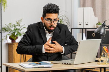 Fototapeta premium Stressed Indian businessman freelancer suffers from tension holding chest with heart pain attack risk of stroke symptom while working at modern home office workplace desk. Male Arabian in formal suit
