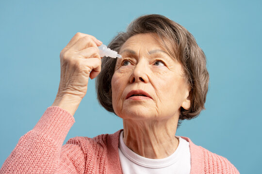 Elderly woman applying eye drops for glaucoma treatment on blue background