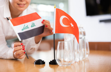 Little flag of Turkey on table with bottles of water and flag of Iraq put next to it by positive young woman in meeting room
