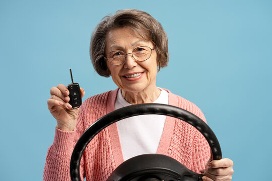 Elderly woman holding car key and steering wheel smiling on blue background - Powered by Adobe