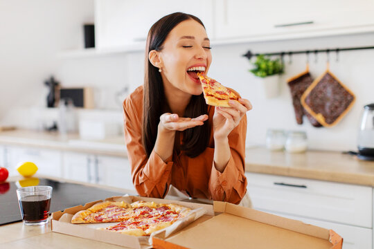 The joy of pizza. Excited woman enjoying tasty pizza, holding and biting slice with closed eyes, sitting at table in kitchen. Cheat meal, delivery service concept