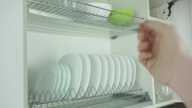 Man takes large plate from dish dryer shelf with crockery and utensils. Man takes white plate from wall cabinet with dishes in light kitchen