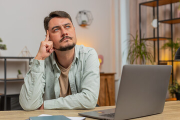 Obraz premium Smart focused man freelancer guy pensive thoughtful sitting at table home office sitting with laptop thinks about solving problem with online service app makes decision deep in thoughts. Lifestyle