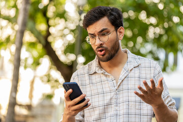 Indian young man using smartphone typing browsing, loses becoming surprised sudden lottery results...