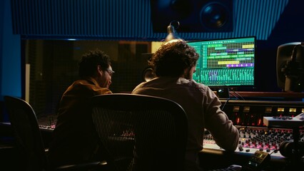 Diverse team of artist and sound designer working on editing tunes for a new hit, operating mixing console with moving faders and colored meters. Musician and producer making tracks. Camera B.