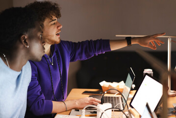 Two young men, one in a purple hoodie and the other in a blue sweater, sit at a desk with laptops and tablets, actively discussing and analyzing data in a dimly lit office.