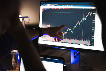 A close-up view of a hand pointing at a computer monitor displaying stock market data and financial charts, with a trading setup in a modern office.