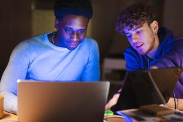 Two young men in a dimly lit office are deeply engaged in analyzing data on a laptop and tablet, with financial charts and a trading setup in the background.