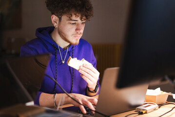 A young man wearing a purple hoodie takes a bite of a sandwich while typing on a laptop at a wooden desk in a modern office with multiple electronic devices.