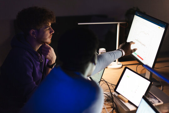 Two young men in a dimly lit office space analyzing stock market data on a large monitor, one actively pointing at the screen while the other listens attentively.