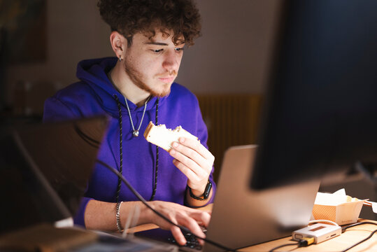 A young man wearing a purple hoodie takes a bite of a sandwich while typing on a laptop at a wooden desk in a modern office with multiple electronic devices.