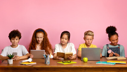 Multiethnic children with electronic devices and books studying at table over pink background, free space. Panorama