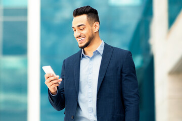Fototapeta premium Happy middle-eastern entrepreneur checking emails on smartphone, having break after business meeting, walking by business center outdoors, arab guy businessman with cellphone and laptop, copy space