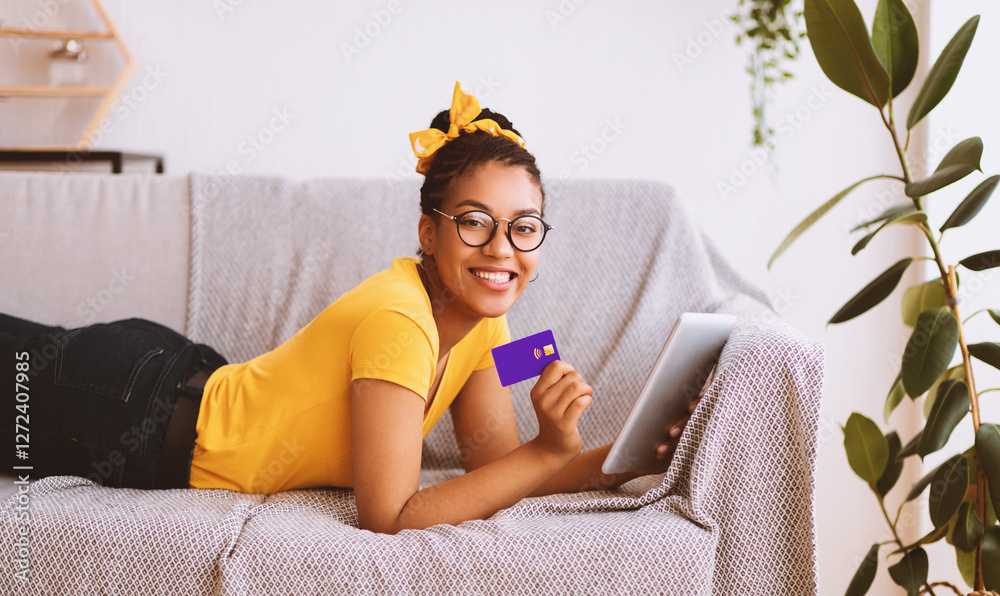 Poster Easy Online Payment Concept. Smiling black lady in glasses using digital tablet and showing credit card, ordering purchases via internet. Woman lying on the couch and looking at camera