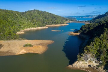 Naklejka premium Aerial view of a receding reservoir, revealing exposed shoreline and surrounding lush forest. Low water levels expose the lakebed. Lower Huia Dam, Auckland, New Zealand