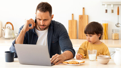Business from home. Young concentrated man consulting clients with cellphone and laptop on weekends, his cute little son eating cookies with milk nearby, sitting at kitchen, free space