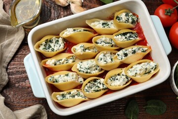 Delicious conchiglie pasta with ricotta cheese, spinach and tomato sauce in baking dish on wooden table, flat lay