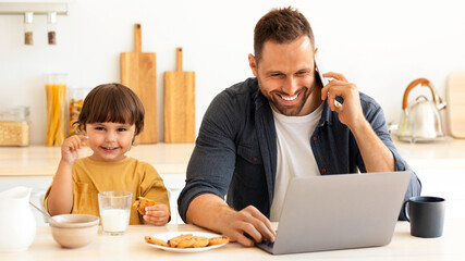 Positive family weekends. Cute little boy with glass of fresh milk smiling to camera, sitting at kitchen with his positive father freelancer working from home with laptop and smartphone