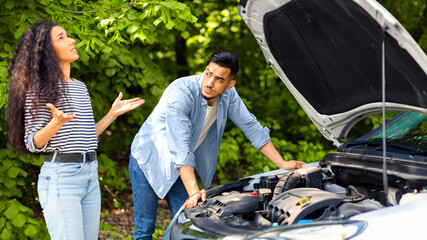 Upset middle-eastern couple having broken car while countryside trip, puzzled arab guy checking engine, emotional brunette lady looking up, sad mixed race family stuck in the middle of road