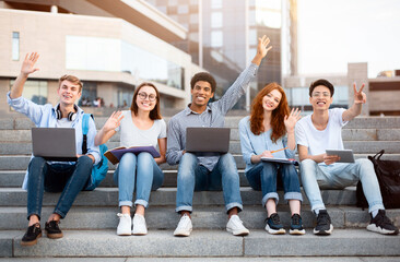 Happy Learning. Diverse group of teenagers waving and raising hands up in the air, sitting on steps with laptops and books