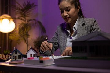 A woman is sitting at a desk with two model houses in front of her. She is holding a pencil and she...