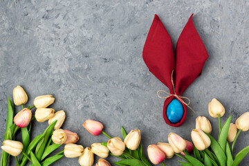A blue Easter egg and a red napkin as bunny or rabbit ears. Easter celebration concept. Top view vertical photo of colorful easter eggs and flowers on grey background