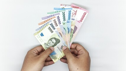 two hands holding a collection of Indonesian banknotes in denominations 1000, 2000, 5000, 10000, 20000, 50000, 75000, 10000 rupiah