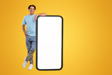 Full body length of smiling casual guy standing and leaning on huge giant cell phone with empty white screen template, orange studio background. Mock up for app website, free space for advertising
