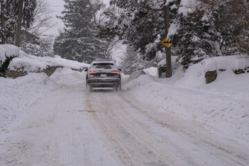 SUV vehicle drives on the snow covered residential city road with big snowbanks on the sides during heavy snowstorm in Toronto, Ontario, Canada. Severe weather and unsafe driving condition concept.