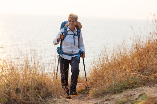 A senior woman with a backpack and trekking poles hikes along a coastal trail surrounded by dry grass. She enjoys the fresh air and scenic ocean view, embracing adventure and outdoor exploration.