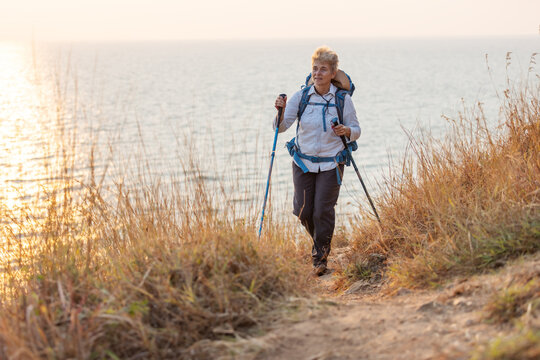 A senior woman with a backpack and trekking poles hikes along a coastal trail surrounded by dry grass. She enjoys the fresh air and scenic ocean view, embracing adventure and outdoor exploration.