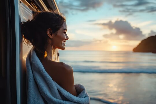 A woman in a towel standing by the camper van, smiling and looking out at the ocean after a refreshing swim, with the beach and sky glowing in the warm light.