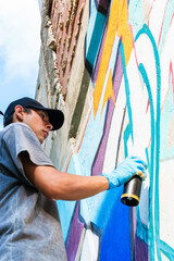 Young graffiti artist wearing a baseball cap and gloves adding vibrant colors to a mural on a weathered urban wall