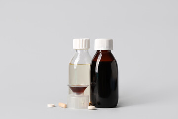 Bottles and measuring cup of cough syrup with different pills on grey background