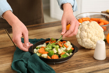 Young woman adding rosemary into frying pan with roasted vegetables in kitchen