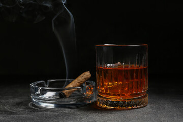 Glass of cold rum and ash tray with cigar on grey grunge background