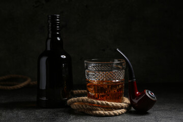 Composition with bottle, glass of cold rum, smoking pipe and rope on black grunge background