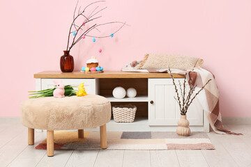 Interior of living room with Easter decorations on commode and flowers at pouf near pink wall
