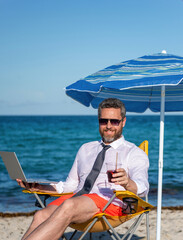Fototapeta premium Businessman on summer. Online freelance work. Businessman worker. Business man using laptop in beach. Travel and business. Man freelance worker online at sea beach. Work-life balance on trips