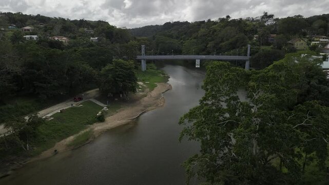 Drone lowers over Macal River facing metal suspension Hawkesworth Bridge with large green tree in the foreground on cloudy day in San Ignacio, Belize