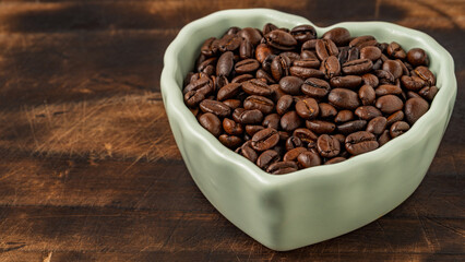 Heart-shaped ceramic bowl filled with roasted coffee beans on a rustic wooden background. Concept of love for coffee and caffeine passion.