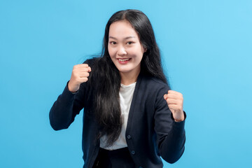 Happy young Asian woman celebrating success with raised fists, wearing casual outfit, standing against blue background. Concept of achievement, victory, motivation, and positive energy.