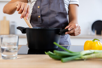 Woman chef using wooden spoon while stirring soup in a pot on induction stove, with fresh...