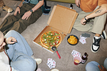 Group of friends relaxing around coffee table, enjoying pizza and snacks, playing card games. Individuals wearing casual attire, creating cozy and fun atmosphere