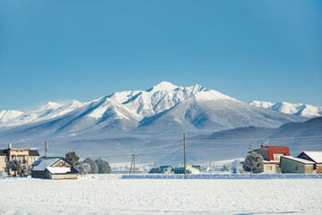 Beautiful winter landscape in Furano, Japan, featuring snow-covered fields, traditional countryside houses, and majestic mountains under a clear blue sky.
