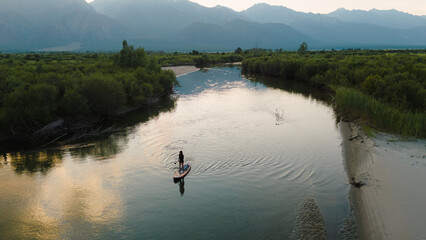 A girl paddleboarding on a serene river surrounded by towering mountains, captured in stunning...