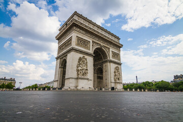 The Arc de Triomphe, seen from a low-angle perspective, dominates the scene under a partly cloudy sky. The cobblestone foreground adds depth, while the Eiffel Tower subtly appears in the background