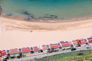 Beachfront houses line the coast while visitors walk along the sandy shore on a sunny day aerial view.