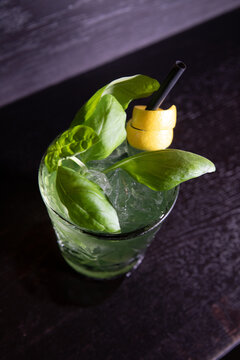 Green cocktail with fresh basil leaves, ice, and a lemon peel garnish, served with a black straw on a dark surface.